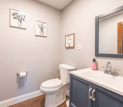 A small bathroom with light gray walls, a white toilet, a white sink with a dark cabinet, a wall mirror, three framed art pieces, wood flooring, and a soap dispenser and flowers on the sink.