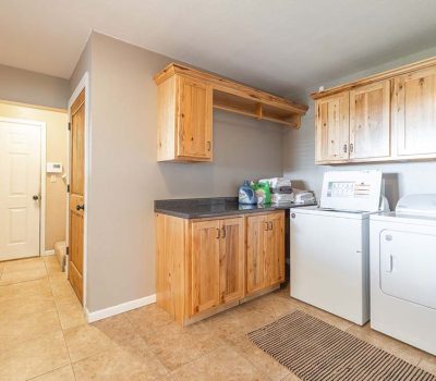A laundry room with light wood cabinets, a countertop with laundry supplies, a washing machine, a dryer, a striped rug on a tiled floor, and a hallway leading to a white door.