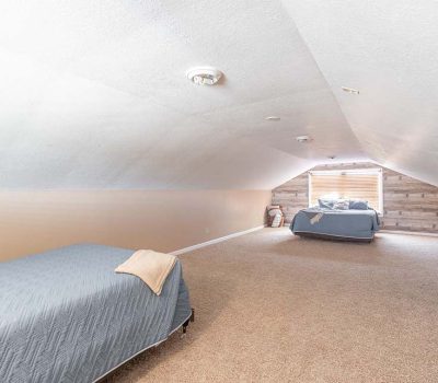 A cozy attic bedroom with sloped ceilings, featuring two beds covered with gray bedding, beige carpeted floor, and a window at the far end letting in natural light.