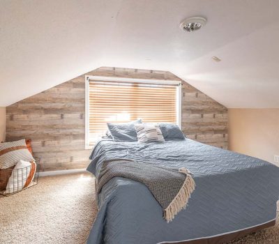 A cozy attic bedroom with sloped ceilings, a large bed covered in blue bedding, a window with wooden blinds behind the bed, and a basket with pillows and blanket on the carpeted floor.