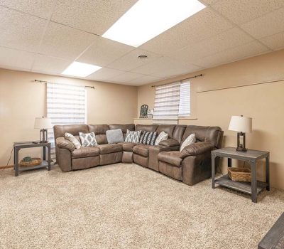 A cozy basement living room with beige walls, carpeted floor, brown sectional sofa, two side tables with lamps, and windows with blinds letting in natural light. A fire extinguisher is mounted on the wall.