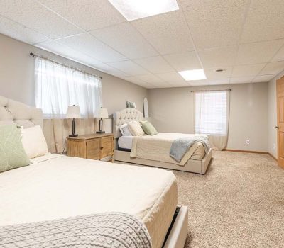Spacious bedroom with two large beds, beige tufted headboards, soft bedding, a nightstand with a lamp, carpeted floor, neutral walls, two windows with curtains, and wooden double closet doors.