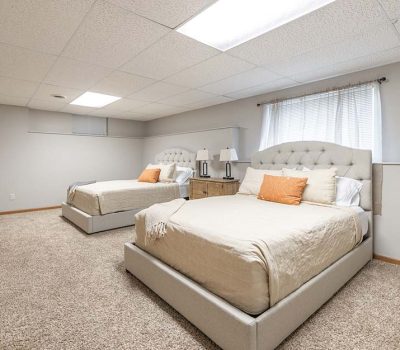 A spacious bedroom with two beige upholstered beds, each with white and orange pillows. A wooden nightstand with lamps sits between the beds. The room has carpeted flooring and a large window with sheer curtains.