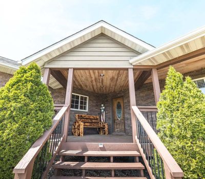 A wooden porch with a bench, flanked by evergreen shrubs, leads to a brown front door under a sloped roof. The house exterior features stone and wood siding.
