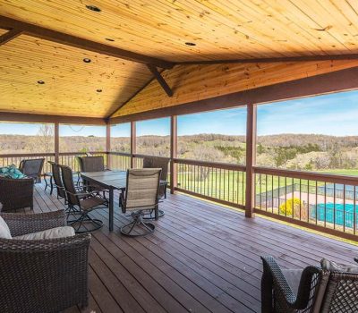 A covered wooden porch with wicker furniture overlooks a green landscape and a blue swimming pool, offering a scenic view of trees and fields under a clear sky.