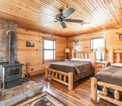 A cozy log cabin bedroom with two wooden beds, a wood-burning stove on a stone hearth, wooden walls and ceiling, ceiling fan, lamps, and windows letting in natural light.