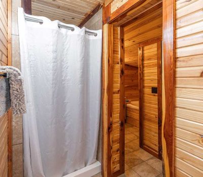 A small bathroom with wood-paneled walls and ceiling, featuring a shower with a white curtain, a towel hanging on a rack, and a wooden doorway leading to a tiled hallway.