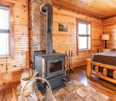A cozy log cabin room with a wood-burning stove on a stone hearth, surrounded by wood-paneled walls, a log-framed bed, and windows letting in natural light. A wicker basket with firewood sits by the stove.