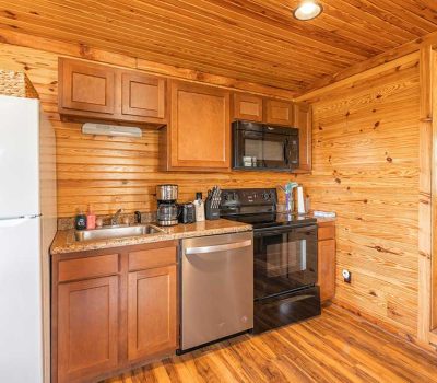 Small rustic kitchen with wooden walls and ceiling, featuring a refrigerator, sink, dishwasher, stove, microwave, coffee maker, and countertop appliances. An open doorway leads to a cozy room with visible windows.