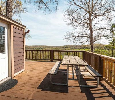 A wooden deck with a picnic table overlooks a scenic, tree-filled landscape. The deck is attached to a brown cabin with a white door, and distant rolling hills are visible under a clear sky.