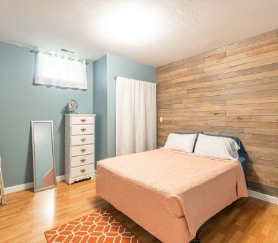 Cozy bedroom with a wood accent wall, a bed with peach bedding, blue walls, a white dresser, a standing mirror, a ladder-style towel rack, and a small orange rug on light wood floors.