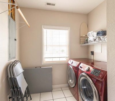 A laundry room with a red washer and dryer, a window with blinds, a shelf holding laundry items, and several folded chairs leaning against the wall on a white tiled floor.