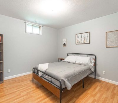 A minimalist bedroom with light gray walls, a metal bed with gray bedding, wood flooring, a bookshelf with books and decor, and three framed pictures on the wall. A small window lets in natural light.