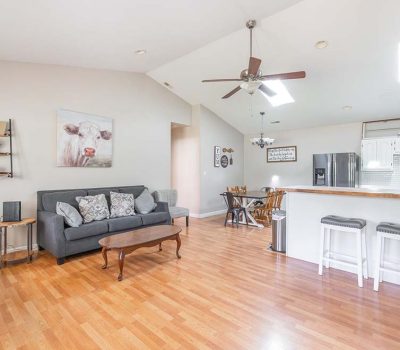 Bright open-concept living area with wood floors, gray couches, a coffee table, wall art, a ceiling fan, dining table, and a kitchen with white cabinets, stainless appliances, and two barstools at the counter.