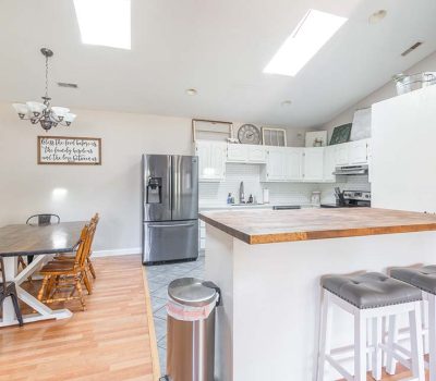 Bright kitchen and dining area with white cabinets, stainless steel appliances, a wooden dining table with chairs, two bar stools at a wooden counter, and natural light from skylights and windows.
