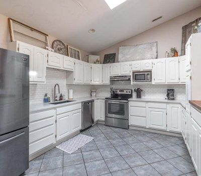 A spacious kitchen with white cabinets, stainless steel appliances, gray tile floor, and wood countertops. There is a refrigerator on the left, an oven and microwave in the center, and kitchen decor on the upper shelves.