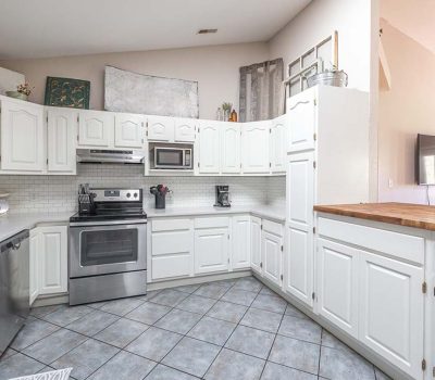 Bright, modern kitchen with white cabinets, stainless steel appliances, tiled floor, and a wooden countertop island. Artwork and decor are displayed above the cabinets, and a TV is visible in the adjacent room.