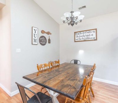 A dining area with a wooden table, six mixed chairs, wall art with food-themed quotes, a chandelier above, and a stainless steel refrigerator on light wood floors.