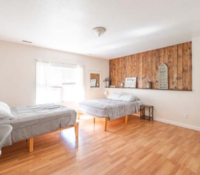 Bright bedroom with two beds, light wood flooring, a wooden accent wall, simple decor, a window with sheer curtains, and a small desk with a chair. The room appears clean, spacious, and minimally furnished.