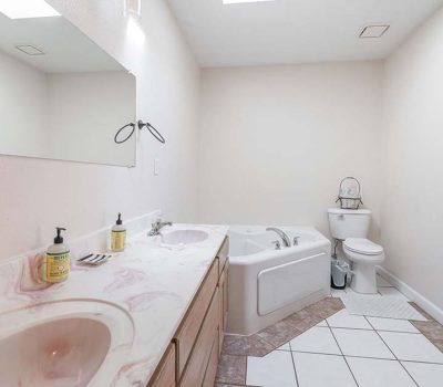 Bathroom with a double sink vanity, large mirror, corner bathtub, and toilet. The countertop has soap dispensers and toothbrushes, and the floor is a mix of white and brown tiles. The room is lit by natural light.
