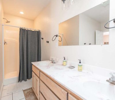 A bright bathroom with a double sink vanity, two mirrors, soap dispensers, and a shower with a dark curtain. The walls are light-colored and the floor has white and brown tiles.