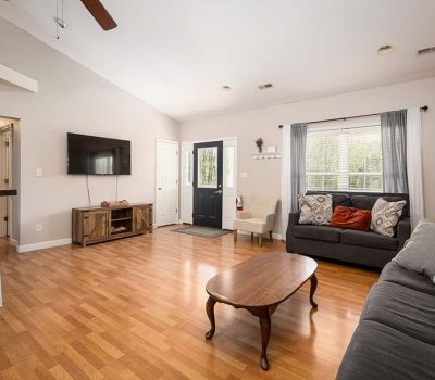 A cozy living room with wood floors, two gray sofas with patterned pillows, a wooden coffee table, a wall-mounted TV, and large windows with sheer curtains letting in natural light.