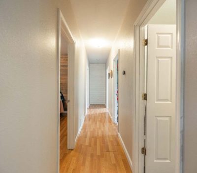 A bright hallway with light-colored walls and wood flooring, featuring several open doors on both sides and lit by ceiling lights, leading to a closed door at the end.