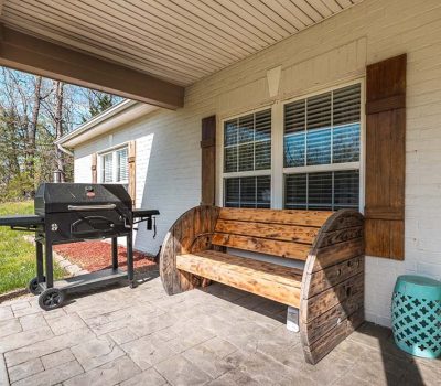 A wooden bench and a barbecue grill sit on a covered patio with stone flooring beside a white brick house; a small blue stool and trees are visible in the background.