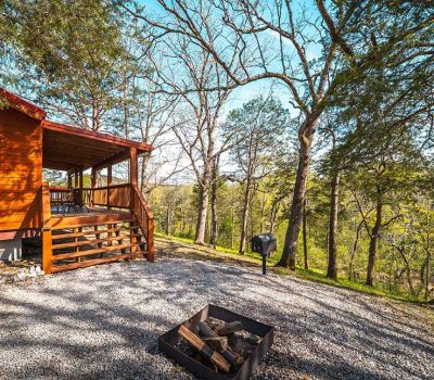 A wooden cabin with a covered porch sits in a forested area. A gravel area with a fire pit filled with logs and a barbecue grill is in the foreground. Trees with sparse leaves surround the scene under a blue sky.
