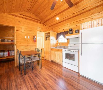 A small cabin kitchen with wood-paneled walls and ceiling, a white refrigerator, stove, and sink. A dining table with four chairs is in the center, and a doorway reveals bunk beds in a cozy bedroom.