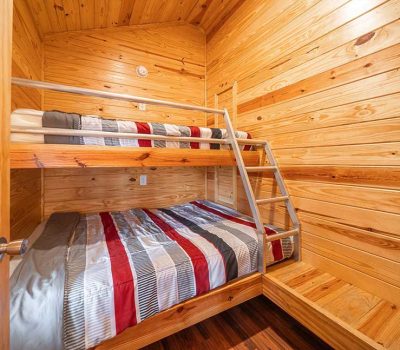 A small wooden room with a bunk bed; the lower bed is full-size and the upper bed is twin-size. Both beds are neatly made with striped gray, white, red, and black bedding. A metal ladder leads to the top bunk.