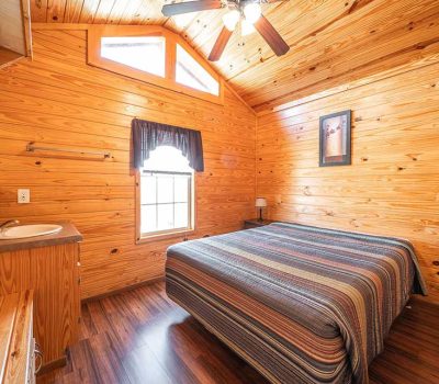 Cozy bedroom with wooden walls and ceiling, a large bed with a striped comforter, a small window with a dark curtain, a ceiling fan, a wall-mounted sink, and a landscape painting above the bed.