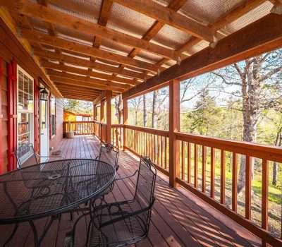 A wooden porch with a covered roof, black metal table and chairs, red window shutters, and a view of trees and greenery in the background on a bright, sunny day.