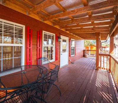 A spacious wooden porch with a metal table and chairs, red siding, large windows with white frames, and a clear roof, overlooking a sunny outdoor area with trees.