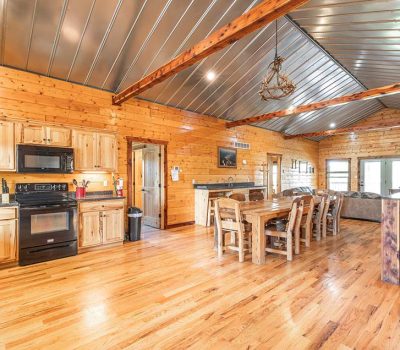 Open-concept log cabin interior with wooden floors and walls, a kitchen area with black appliances, a long dining table with chairs, and a living area with sofas and a stone fireplace. Exposed metal ceiling.