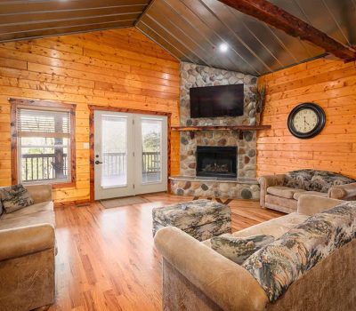 Cozy log cabin living room with wood walls and ceiling, stone fireplace, mounted TV, large clock, three patterned sofas, and glass doors leading to a porch. Warm natural lighting fills the space.