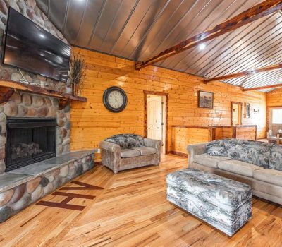 Cozy cabin living room with wood-paneled walls, stone fireplace, wall-mounted TV, plush couches, ottoman, and rustic decor. Exposed beams and hardwood floors create a warm, inviting atmosphere.