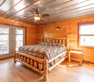 A cozy bedroom with wood-paneled walls and ceiling, a rustic log bed with patterned bedding, a wooden nightstand, ceiling fan, large windows, and glass double doors opening to a deck. Sunlight fills the room.