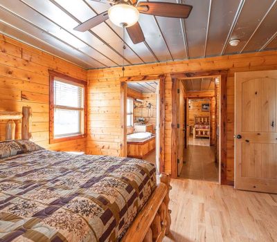 A cozy wooden cabin bedroom with a log-frame bed, patchwork quilt, hardwood floor, ceiling fan, large window, and open doors leading to a bathroom and another room.