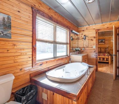 A rustic bathroom with wood-paneled walls, a white toilet, a large bathtub with jets, a window with blinds, a trash can, and a door leading to a bedroom with a wooden bed.