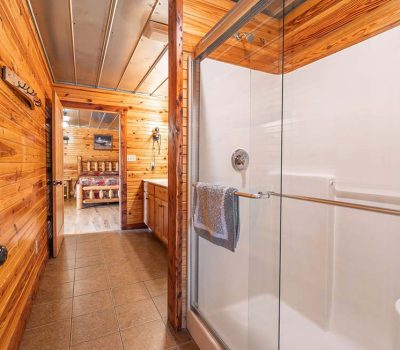 A hallway bathroom with wood-paneled walls, a glass shower with a towel on the handle, and a view into a bedroom with a wooden bed and matching furniture.