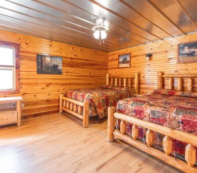 A cozy wooden cabin bedroom with two log-frame beds, colorful patchwork quilts, a wooden desk near a window, and nature-themed artwork on the wood-paneled walls.