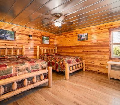 A cozy log cabin bedroom with wooden walls and flooring, featuring two rustic wooden beds with patterned bedspreads, a ceiling fan, wall art, a window, and a small wooden bench.