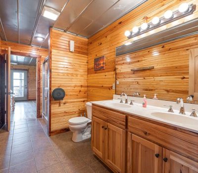 A rustic bathroom with wood-paneled walls, double sink vanity, toilet, large mirror with lights, tiled floor, and an open door leading to a hallway with natural light.