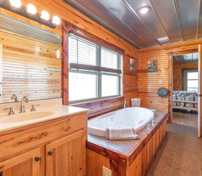A rustic bathroom with wood-paneled walls, a large vanity with a sink and mirror, a jacuzzi-style bathtub with towels, a toilet, and a window letting in natural light. The door is open to a room with a couch.