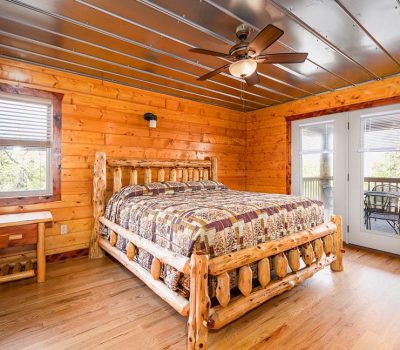 A cozy log cabin bedroom with wooden walls and floors, a rustic wooden bed with a patchwork quilt, a small side table, ceiling fan, and glass doors leading to a balcony with outdoor seating.