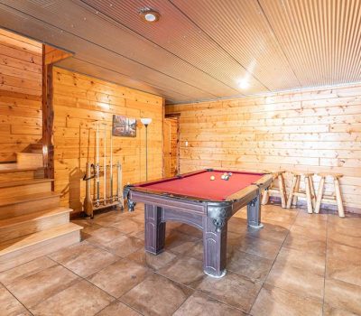 A cozy, wood-paneled game room with tiled floors, a red felt pool table, pool cues on a rack, a stairway, rustic stools, and a floor lamp by the wall.