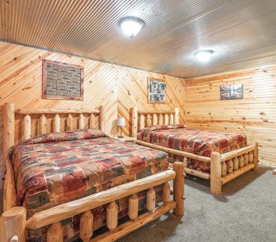 A cozy bedroom with two rustic log-frame beds, colorful patterned bedding, wood-paneled walls and ceiling, gray carpeted floor, a wooden chest, and framed artwork on the walls.