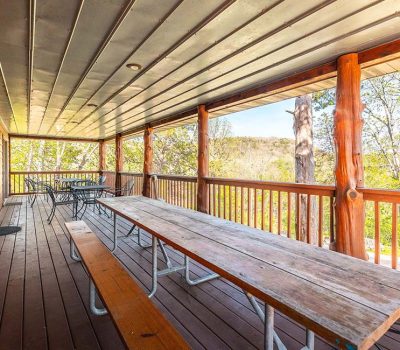 A covered wooden porch with long picnic tables and metal chairs, overlooking a green, tree-filled landscape under a bright sky.