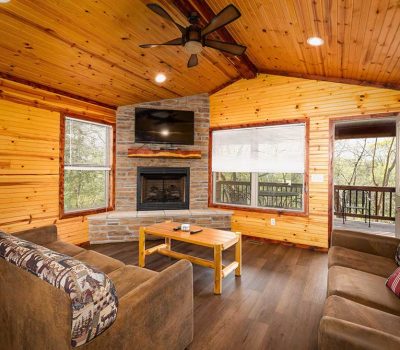 Cozy wooden cabin living room with large windows, a stone fireplace, wall-mounted TV, two patterned sofas, a coffee table, ceiling fan, and an open door leading to a deck with trees visible outside.